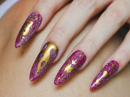 A close-up of a woman’s pointer, middle, ring, and pinky fingers with long, almond-shaped nails. The nails have a purple base with holographic reflective glitter. Gold nail sticker designs decorate each nail: the pointer finger features a snake with tiny diamond-shaped sparkles, the middle finger showcases a flame, the ring finger displays a moth with a moon on top and diamond-shaped sparkles, and the pinky finger has three vertically arranged hearts, with the middle heart being larger than the others.