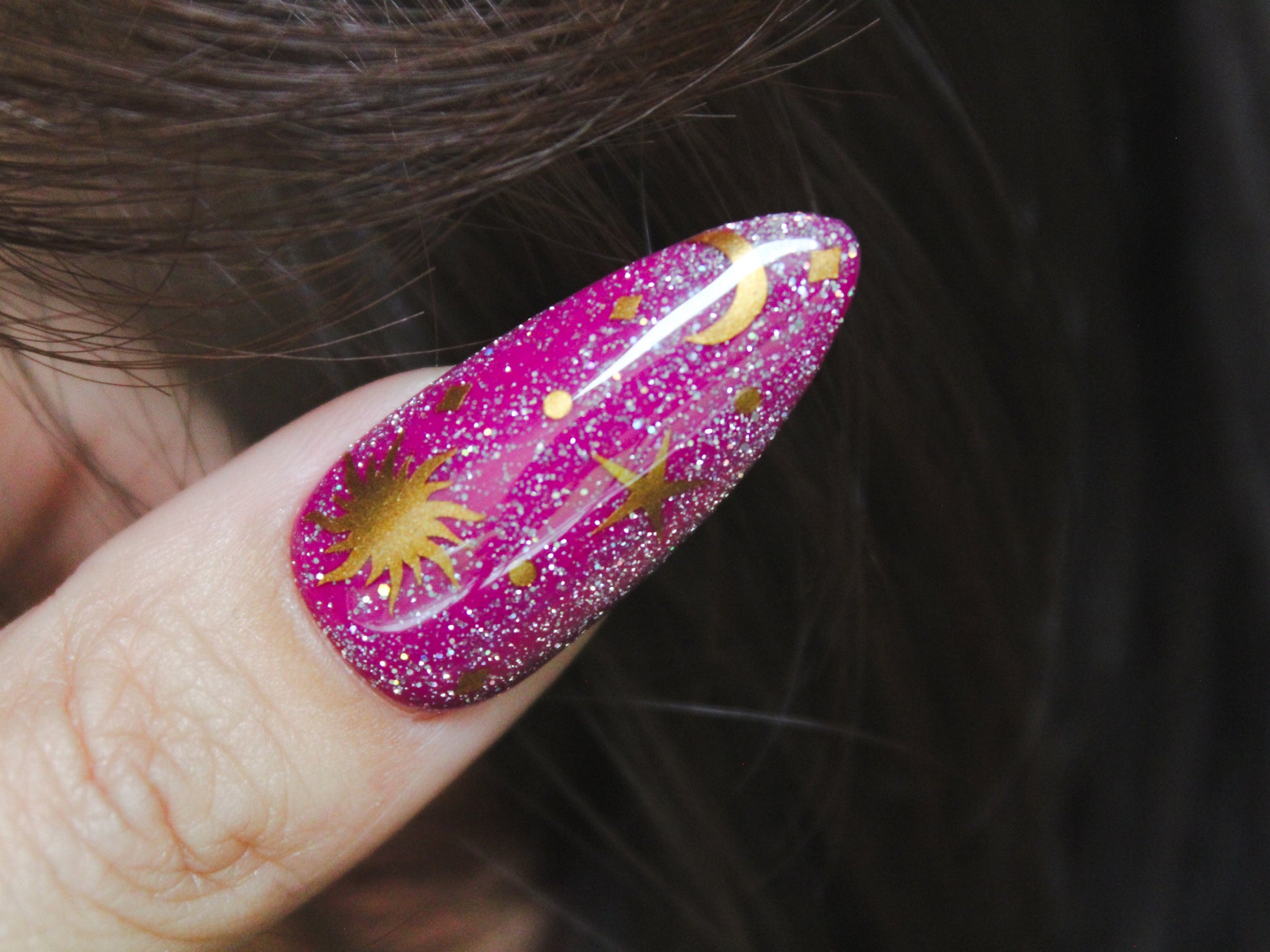 A close-up of a woman’s thumb with a long, almond-shaped nail. The nail has a purple base with holographic reflective glitter. Gold nail sticker designs feature a sun, star, moon, and tiny diamond-shaped sparkles and dots, creating a celestial and elegant look.