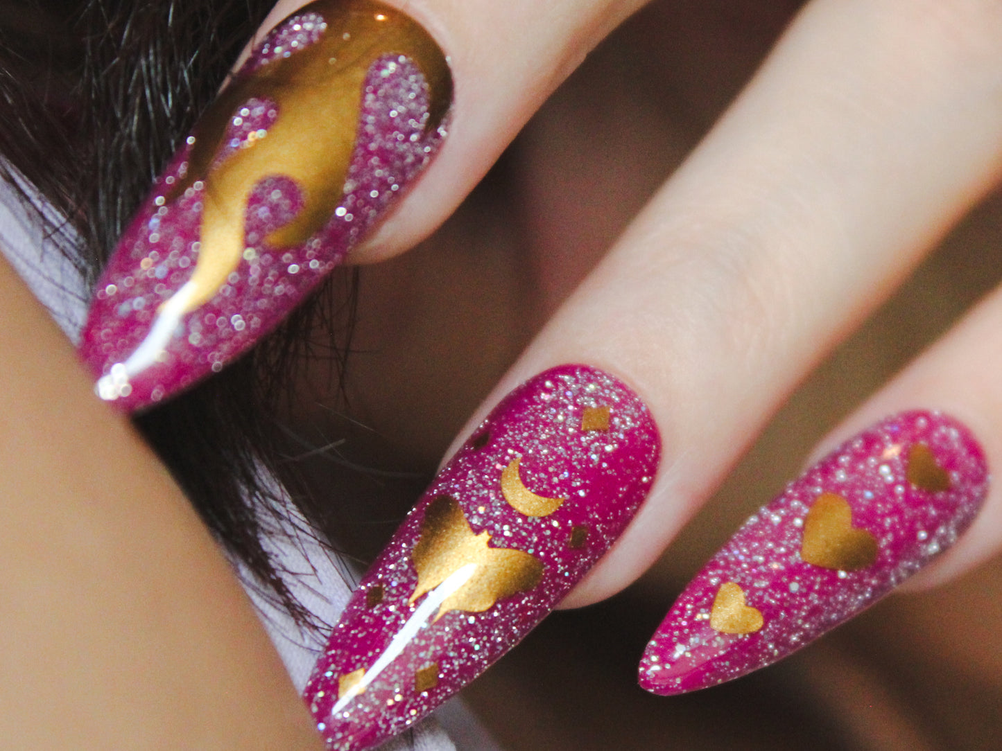 A close-up of a woman’s middle, ring, and pinky fingers with long, almond-shaped nails. The nails have a purple base with holographic reflective glitter. Gold nail sticker designs adorn each nail: the middle finger displays a flame, the ring finger features a moth with a moon on top and tiny diamond-shaped sparkles, and the pinky finger showcases three vertically arranged hearts, with the middle heart being the largest.