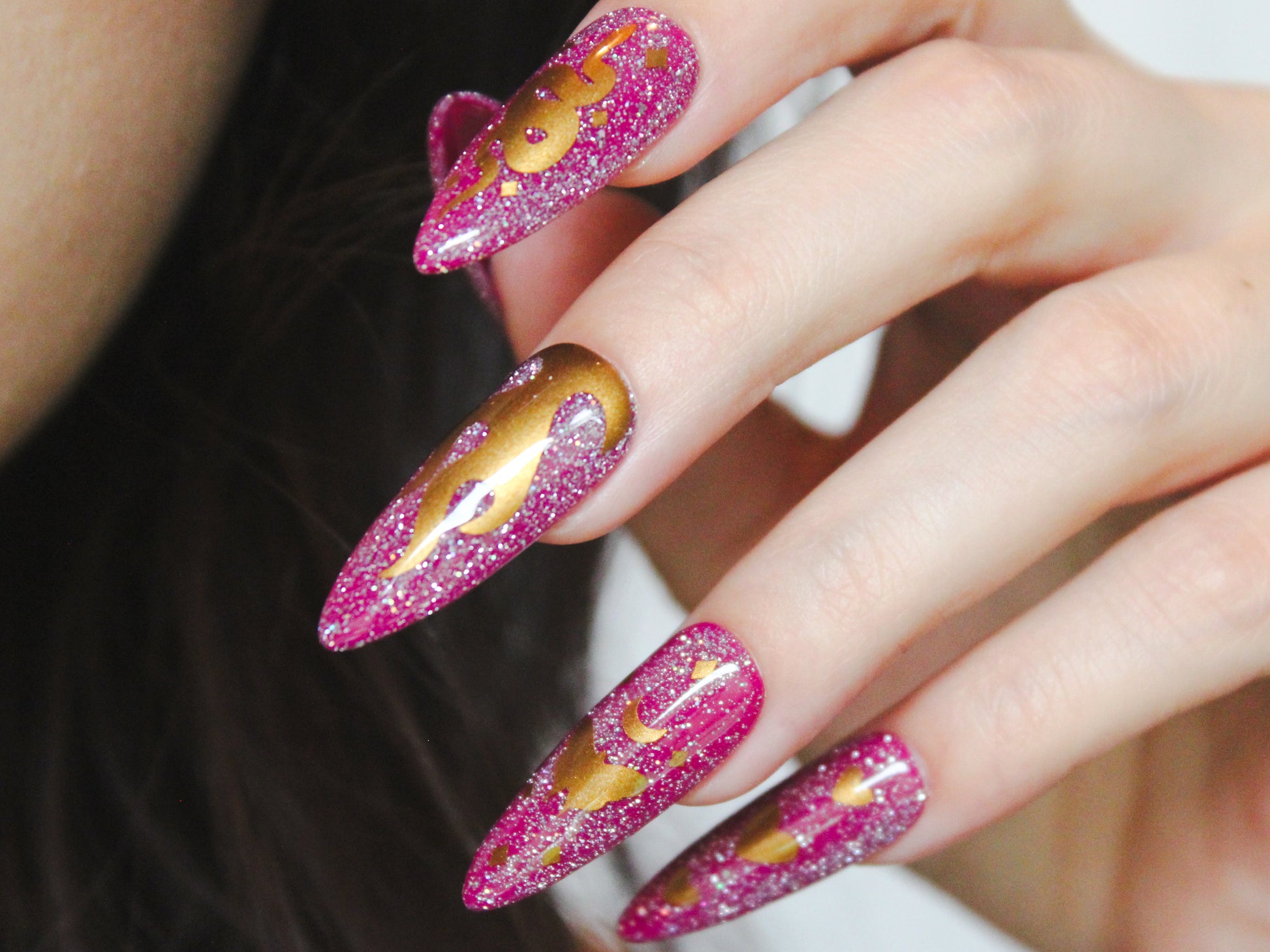 A close-up of a woman’s pointer, middle, ring, and pinky fingers with long, almond-shaped nails. The nails have a purple base with holographic reflective glitter. Gold nail sticker designs decorate each nail: the pointer finger features a snake with tiny diamond-shaped sparkles, the middle finger showcases a flame, the ring finger displays a moth with a moon on top and diamond-shaped sparkles, and the pinky finger has three vertically arranged hearts, with the middle heart being larger than the others.