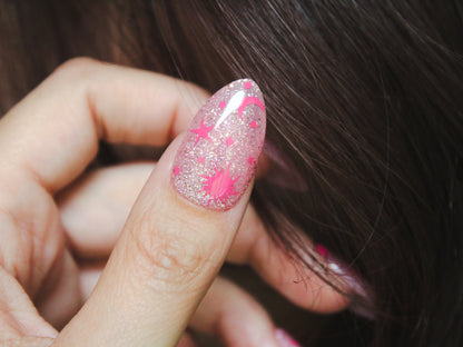 A close-up of a woman’s thumb showcasing ‘Celestial Love Serpent Sparkle Nail Stickers’ in pink. The almond-shaped nail is painted a sheer light pink with a layer of holographic glitter. The medium-length nail features hot pink sticker designs, including a sun, star, moon, and tiny diamond-shaped sparkles and dots, creating a celestial and whimsical look.