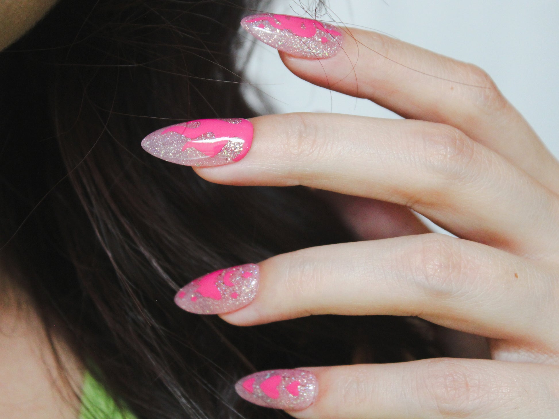  A close-up of a woman’s hand showing her index, middle, ring, and pinky fingers, all with a sheer light pink base and holographic glitter. The nails are almond-shaped and medium-length. The index finger has a pink snake with tiny diamond-shaped sparkles. The middle finger features a flame design. The ring finger displays a pink moth with a moon on top, centered on the nail, with tiny diamond-shaped sparkles. The pinky finger has three pink hearts arranged vertically, with the middle heart being larger than