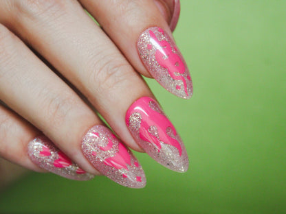 A close-up of a woman’s hand showing her index, middle, ring, and pinky fingers, all with a sheer light pink base and holographic glitter. The nails are almond-shaped and medium-length. The index finger has a pink snake with tiny diamond-shaped sparkles. The middle finger features a flame design. The ring finger displays a pink moth with a moon on top, centered on the nail, with tiny diamond-shaped sparkles. The pinky finger has three pink hearts arranged vertically, with the middle heart being larger than 