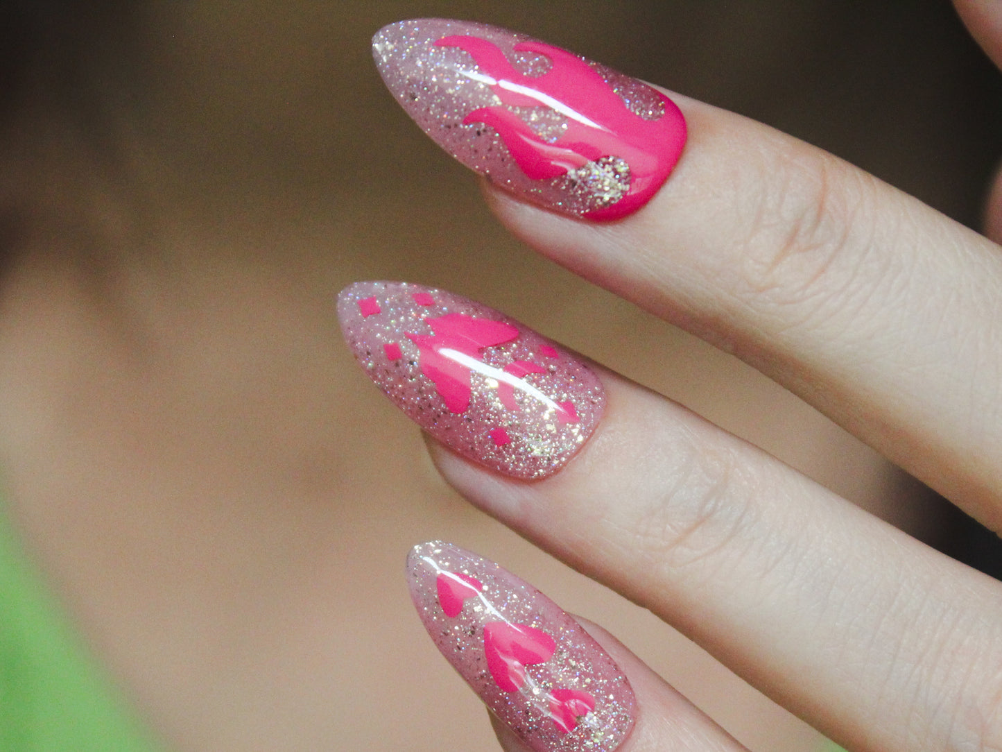 A close-up of a woman’s middle, ring, and pinky fingers with almond-shaped, medium-length nails. The sheer light pink base and holographic glitter accentuate the pink nail sticker designs. The middle finger features a flame, the ring finger showcases a pink moth with a moon on top and tiny diamond-shaped sparkles, and the pinky displays three vertically arranged hearts, with the middle heart being larger than the others.